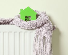 grünes Modellhaus steht auf einem Heizkörper und ist in einen Schal eingewickelt