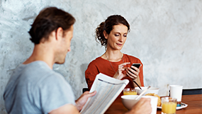 Mann liest Zeitung und Frau benutzt ihr Smartphone beim Frühstück